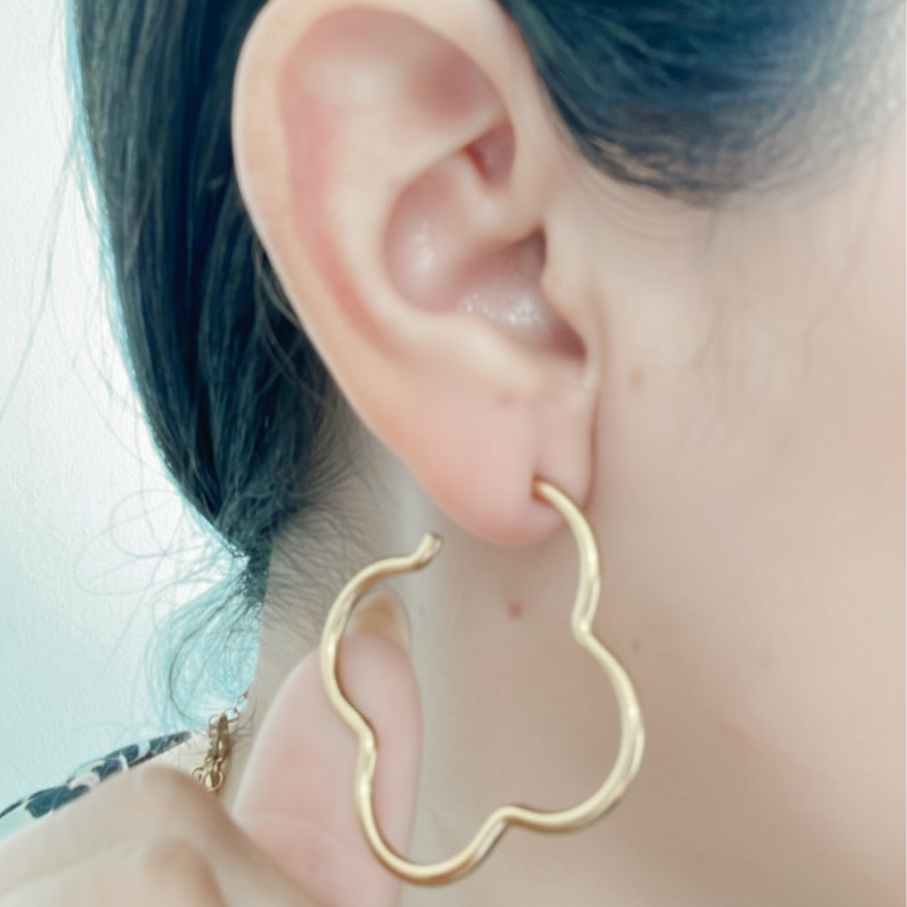 Modelo luciendo aros hoop dorados con forma de trebol en acero inoxidable, estilo moderno de Vanytel Jewelry.