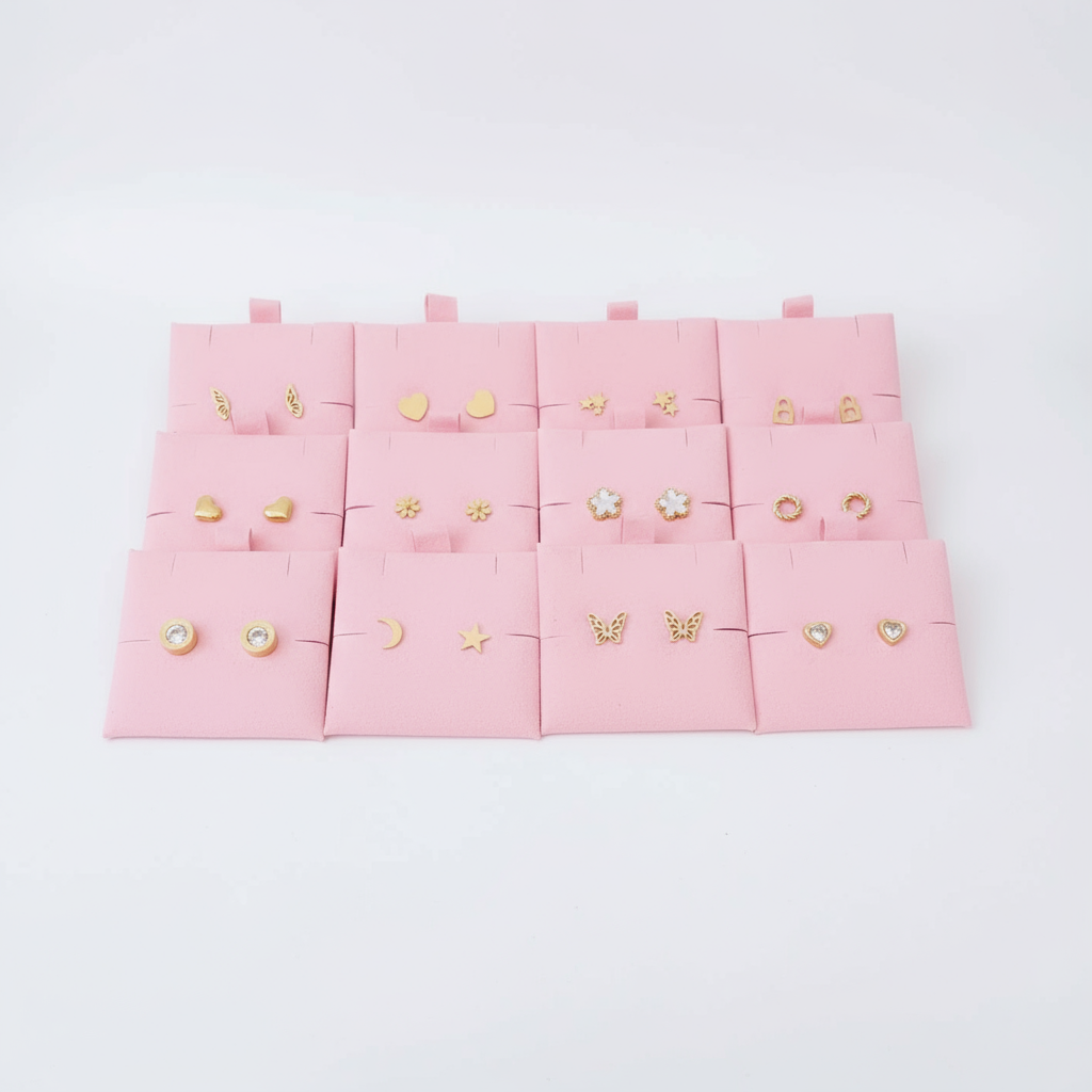 Variedad de aretes dorados tipo stud en acero inoxidable sobre fondo rosa, joyería Vanytel.