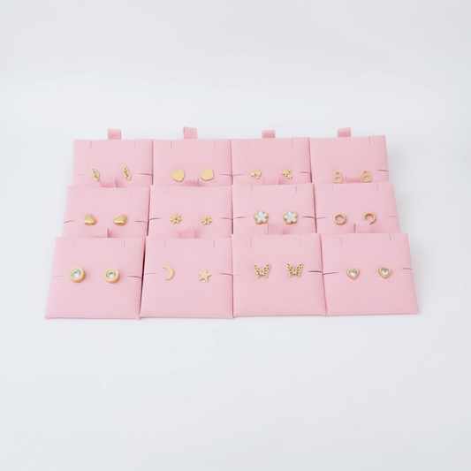 Variedad de aretes dorados tipo stud en acero inoxidable sobre fondo rosa, joyería Vanytel.