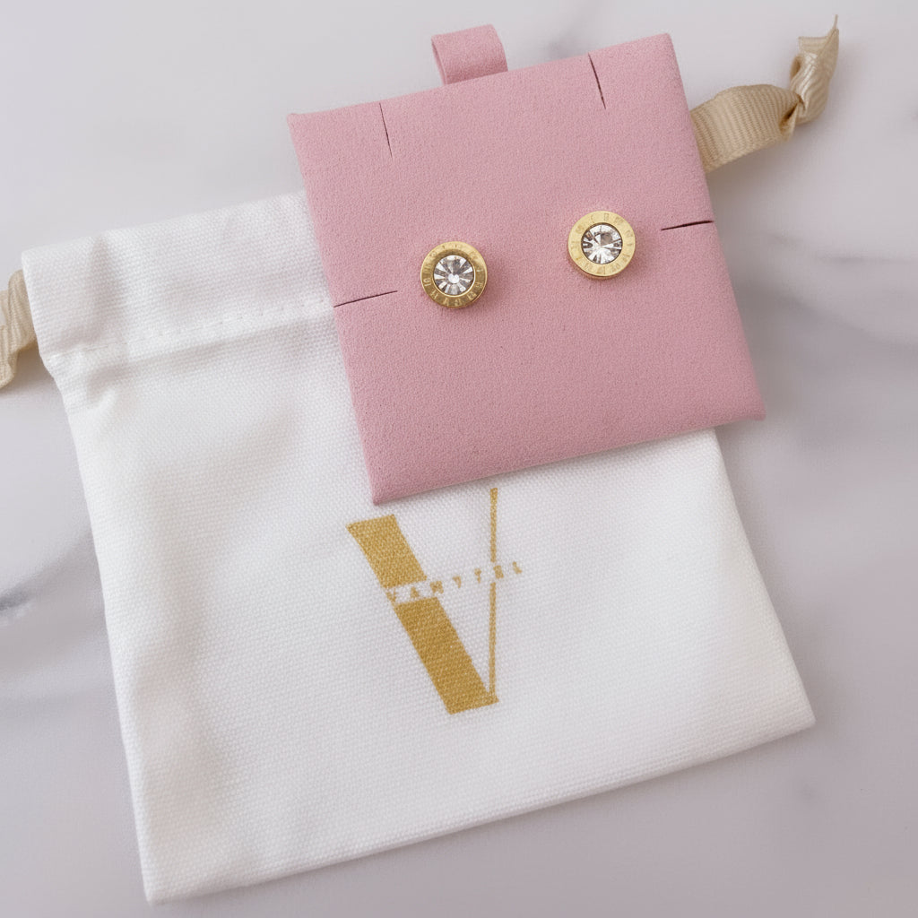 Aretes dorados tipo stud de Vanytel presentados sobre bolsa blanca con logo dorado.
